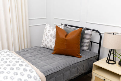 A twin bed made with gray zipper bedding  and an assortment of floral and leather pillows.