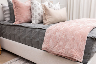 A bed made with gray zipper bedding and decorated with an assortment of floral and pink pillows with a pink velvet blanket folded over the side of the bed.