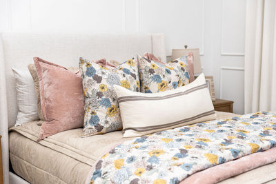 A bed made with tan zipper bedding and an assortment of floral print and pink velvet blankets and pillows.