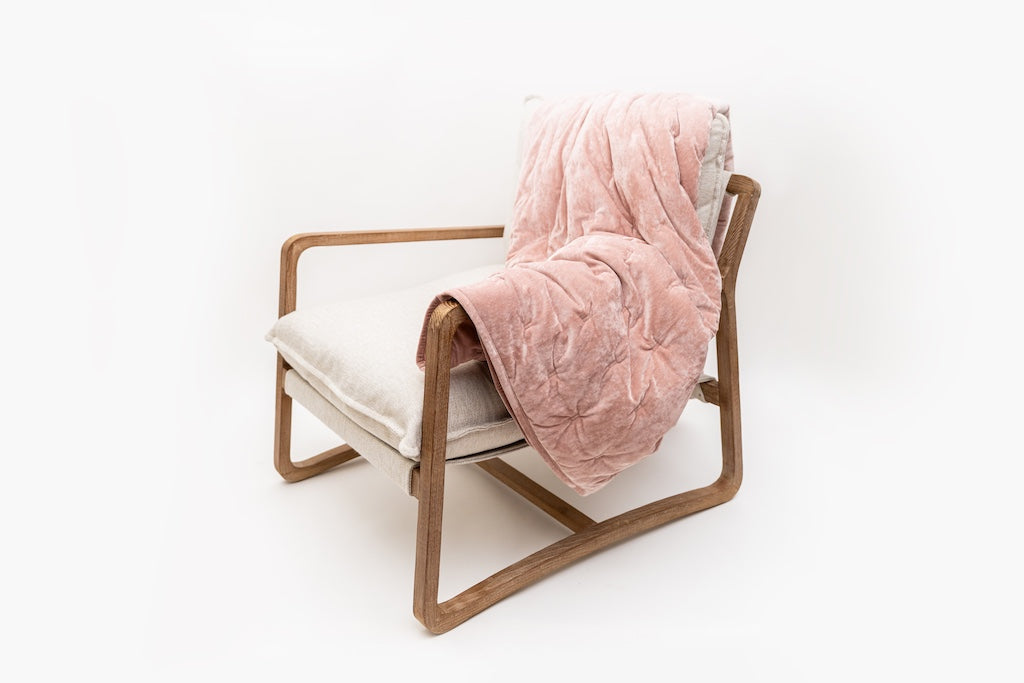 A cushioned chair with a wood frame, a velvet pink blanket draped across the arm and back of the chair.