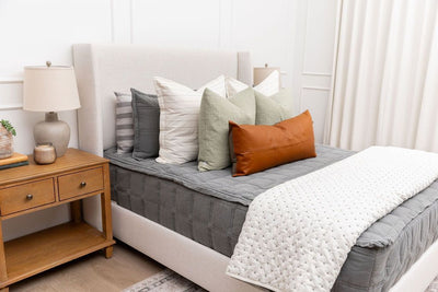 A bed made with gray zipper bedding and an assortment of cream, sage, and leather bedding accessories