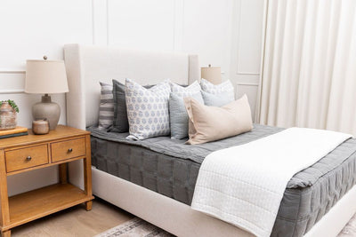 A bed made with gray zipper bedding and an assortment of white and blue floral throw pillows and a white throw blanket at the foot of the bed.