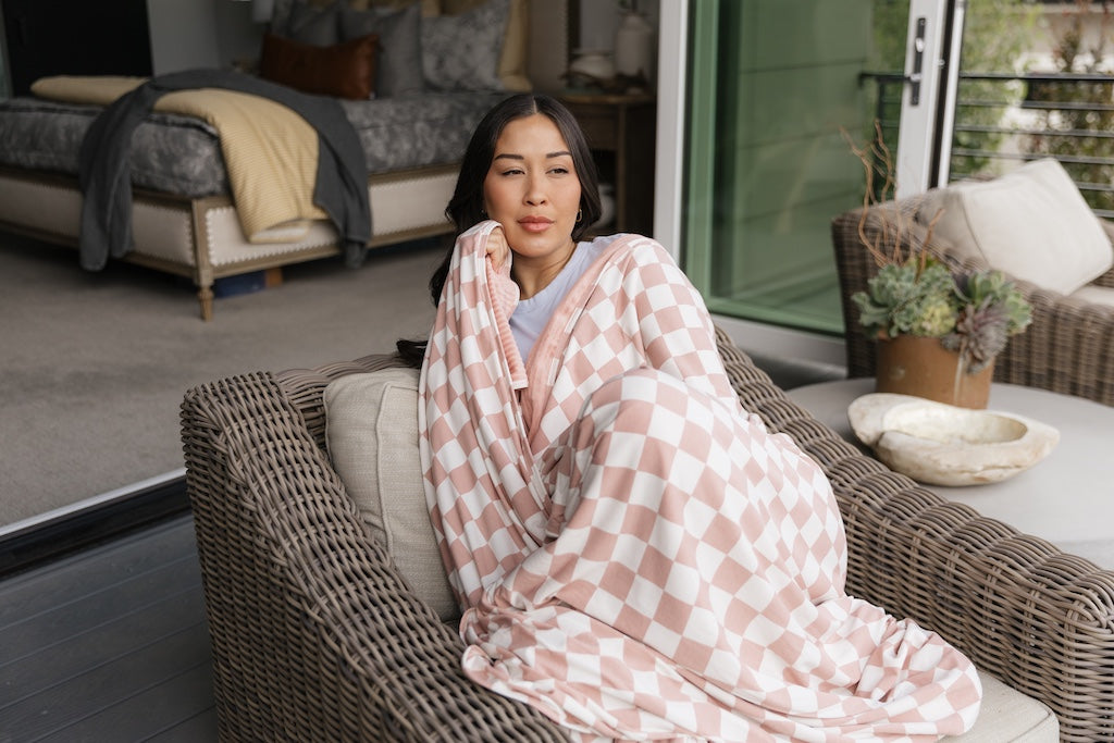 A woman sitting on a chair cuddled up in a pink check travel blanket. 