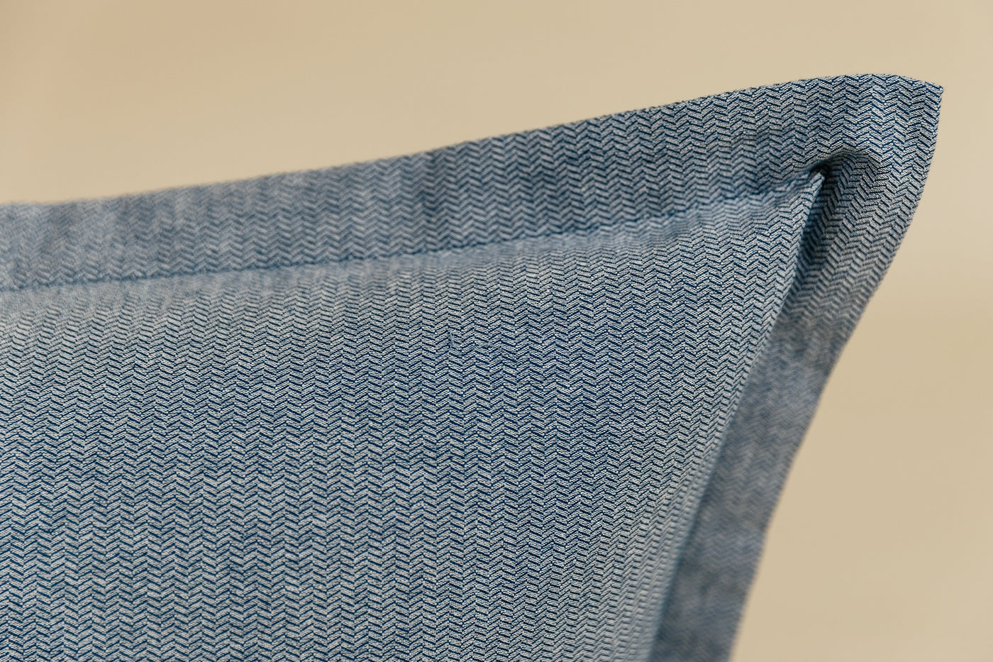A close up corner view of a blue two-toned herringbone euro pillow arranged on a cream background.