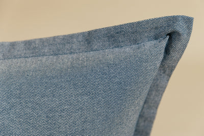 A close up corner view of a blue two-toned herringbone euro pillow arranged on a cream background.