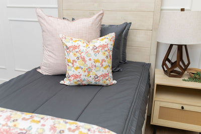 A close up view of a twin bed made with gray zipper bedding and pink and multi-color floral pillows and blanket.