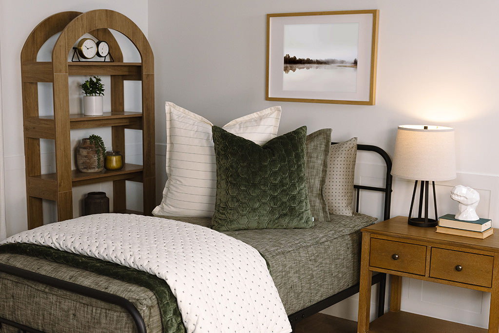 A twin bed made with deep emerald green zipper bedding and an assortment of white and velvet green pillows and blankets.