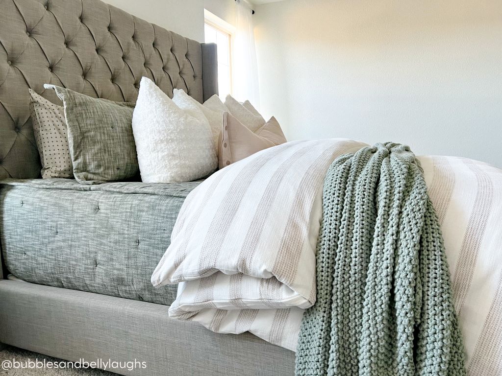 A side view of a bed made with deep emerald green zipper bedding and a tan and white striped duvet with an assortment of neutral pillows and a green woven blanket.