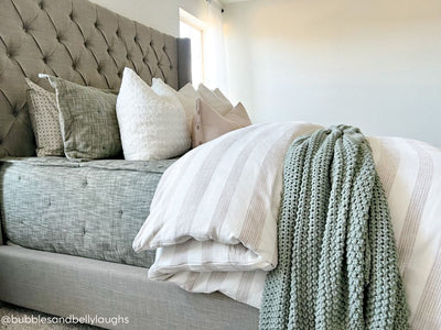 A side view of a bed made with deep emerald green zipper bedding and a tan and white striped duvet with an assortment of neutral pillows and a green woven blanket.