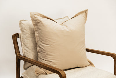 A neutral large tan pillow with frayed edges arranged on a cream fabric chair.
