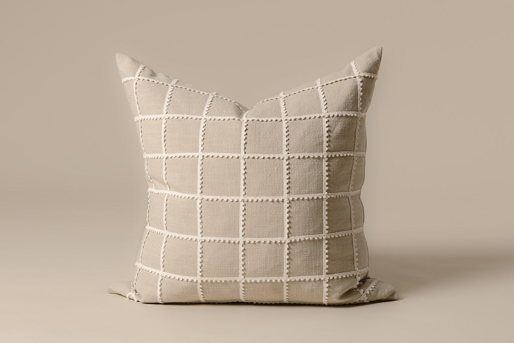 Tan base and a cream trimmed grid pattern detailed pillow on a cream backdrop.