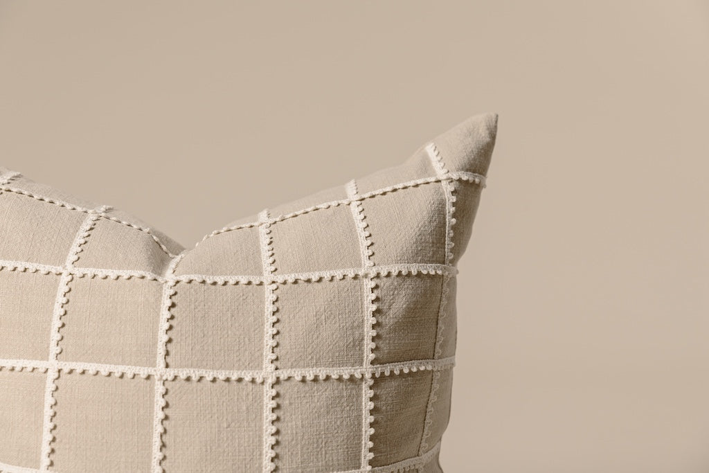Tan base and a cream trimmed grid pattern detailed pillow on a cream backdrop.