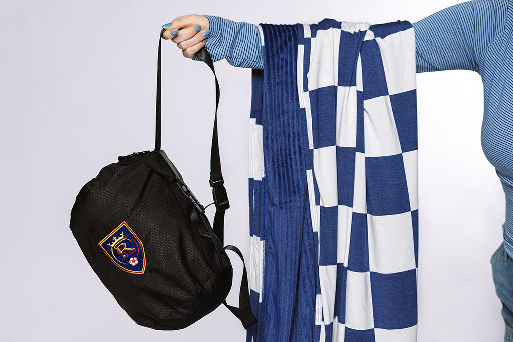 A woman with a blue and white checkered blanket draped over her arm and holding a black bag on a white backdrop.