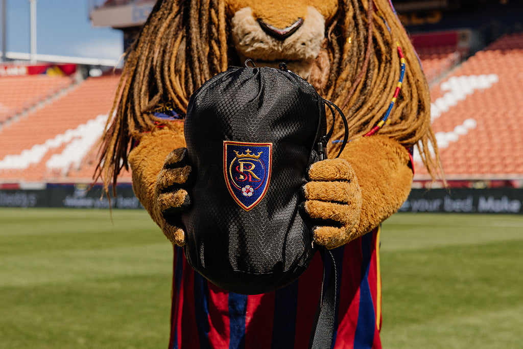 A lion mascot holding a black bag that has a RSL soccer logo on it. 