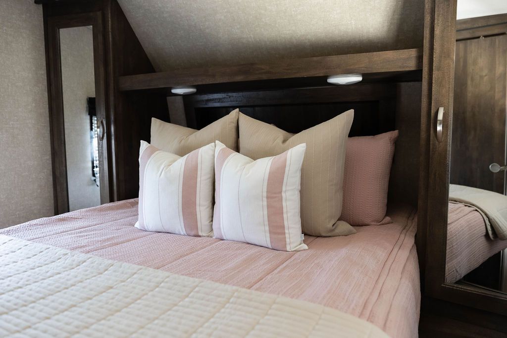 A bed in an RV made with woven pink zipper bedding and an assortment of neutral and pink striped pillows and blanket.