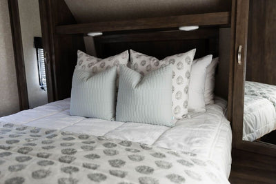A bed in a RV made with white woven plaid zipper bedding and an assortment of blue and gray pattern pillows and blanket.