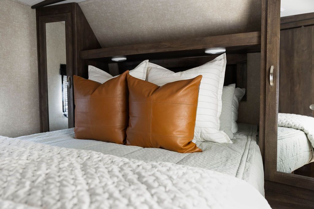 A bed in an RV made with sage green zipper bedding and an assortment of neutral and leather pillows and blanket.