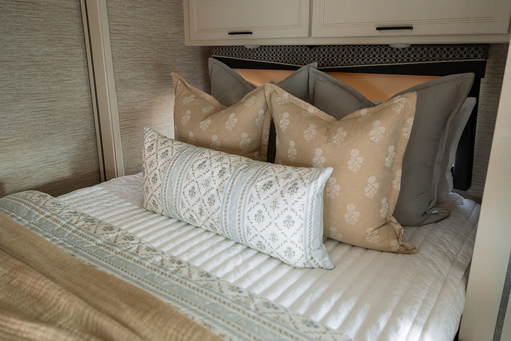 A bed made in an RV with white stripe knitted zipper bedding and an assortment of tan, grey, blue and white floral pillows and blanket.