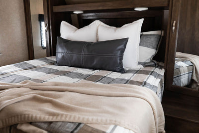 A bed made in an RV with cream, charcoal, and tan plaid zipper bedding and an assortment of neutral pillows and blanket.