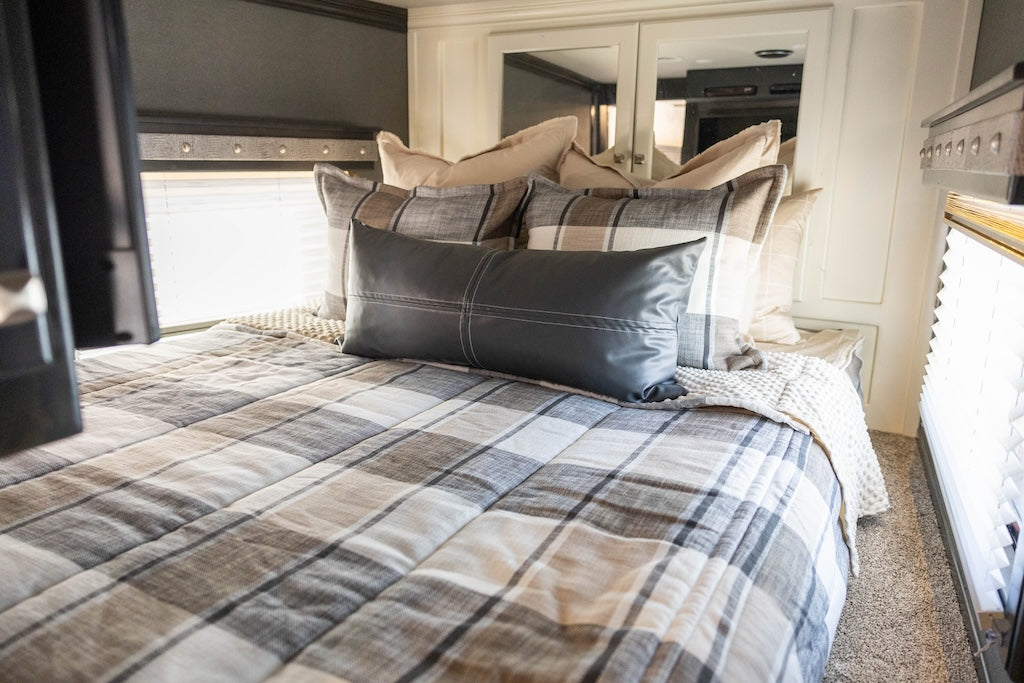 A bed made in an RV with cream, charcoal, and tan plaid zipper bedding and an assortment of neutral pillows.