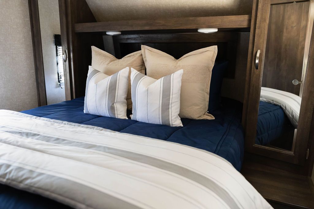 A bed in an RV made with navy blue zipper bedding and an assortment of neutral and gray striped pillows with a blanket at the end of the bed.