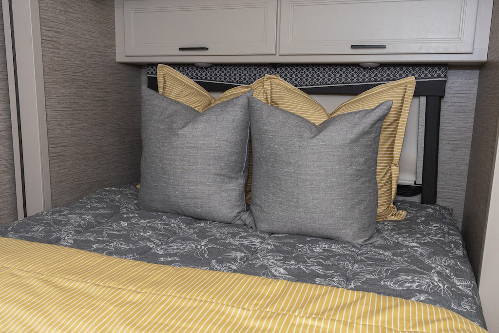 A bed made in an RV with grey and white sketched floral zipper bedding and an assortment of gray and yellow striped pillows and blanket.