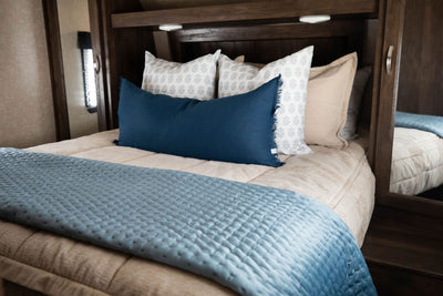 A bed in an RV made with tan faded stripes zipper bedding and an assortment of neutral and blue pattern throw pillows and blanket.