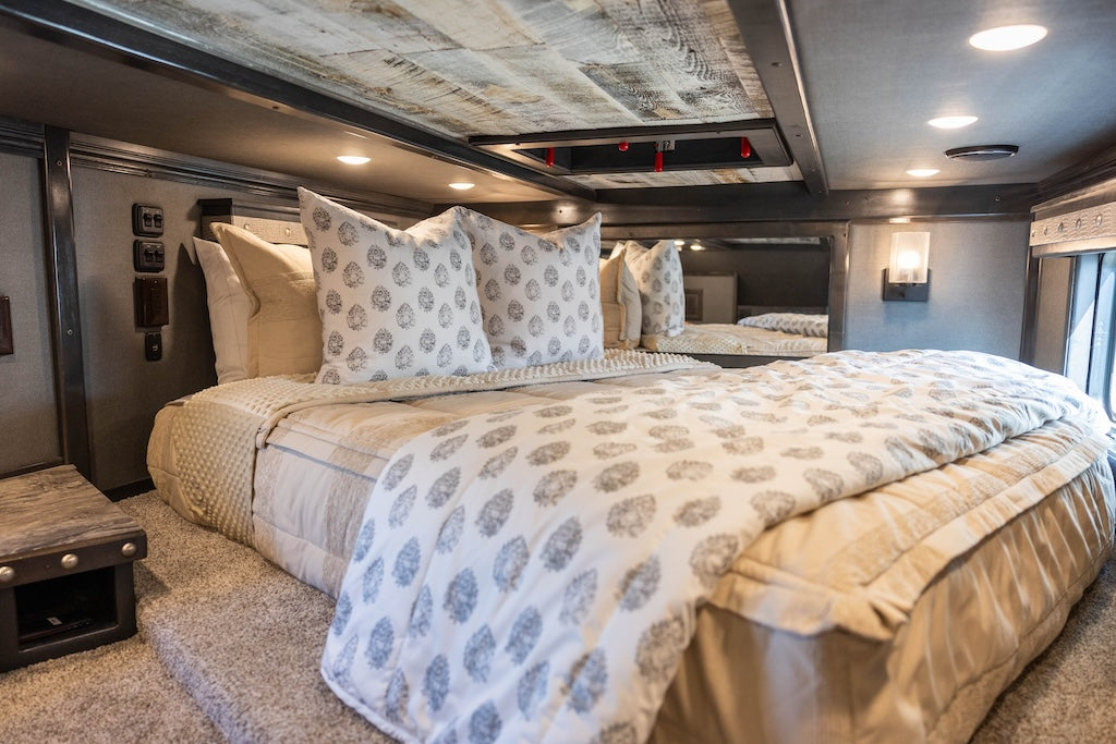 A bed made in an RV tan knitted zipper bedding and an assortment of tan, white and gray floral pillows and blanket.