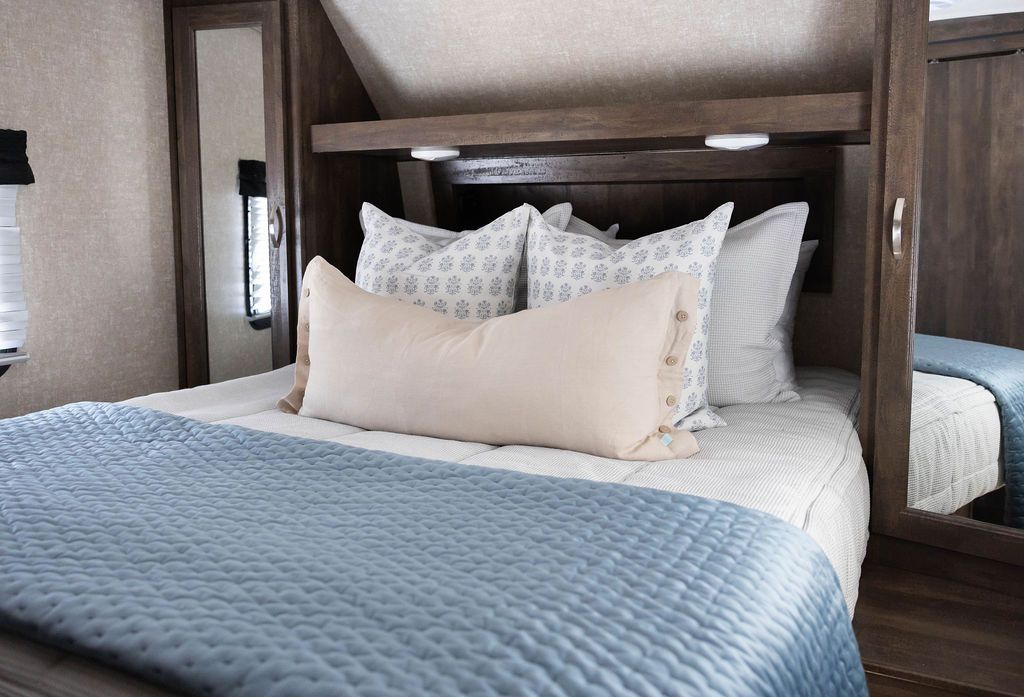 A bed in an RV made with light gray waffle textured zipper bedding and neutral pattern pillows and matching blue velvet blanket.