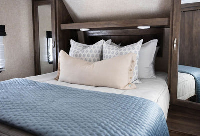 A bed in an RV made with light gray waffle textured zipper bedding and neutral pattern pillows and matching blue velvet blanket.