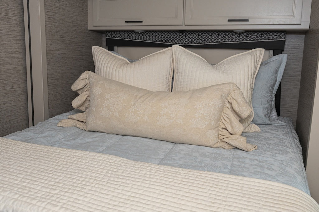 A bed made in an RV with blue floral embossed zipper bedding and an assortment of cream pillows and blanket.