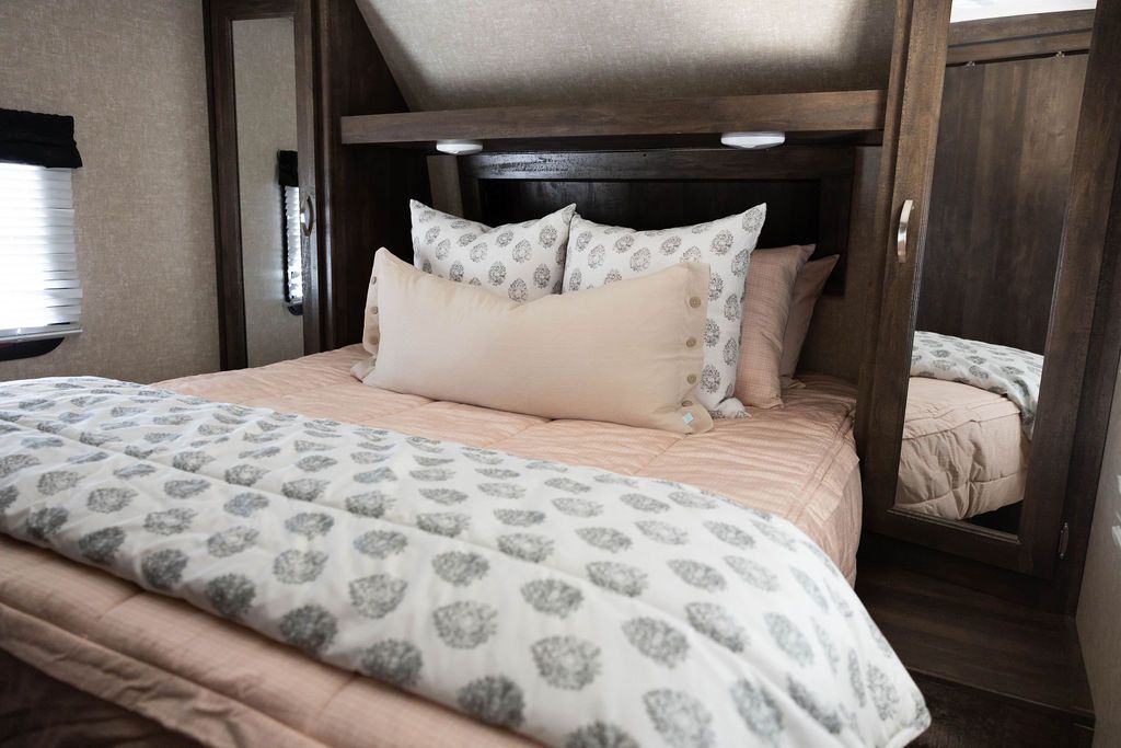 A bed made in an RV with peach pink zipper bedding and adorned with neutral and gray patterned pillows and blanket.