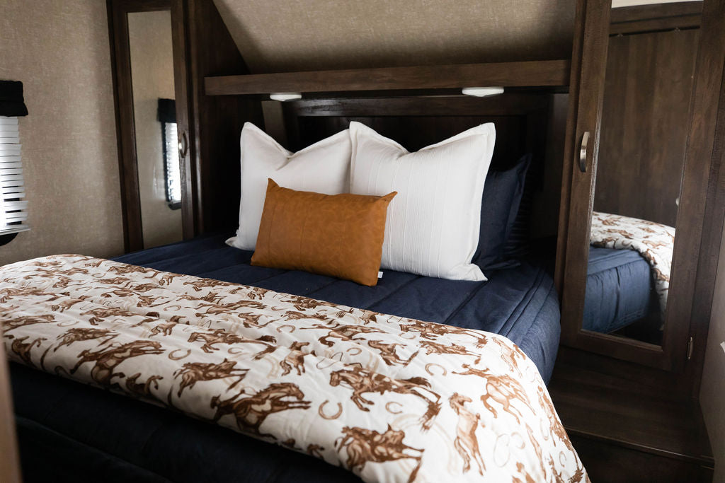 A bed in an RV made with dark denim blue zipper bedding and white and leather pillows and a western printed blanket.