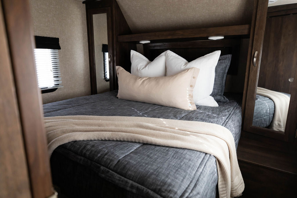 A bed in an RV made with dark charcoal gray zipper bedding and white and tan throw pillows and matching woven blanket.
