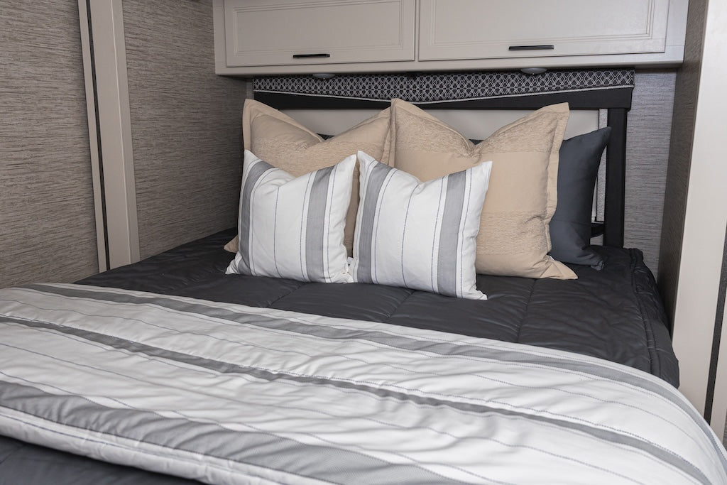 A bed made in an RV with gray zipper bedding and an assortment of tan, white and gray pillows and blanket.