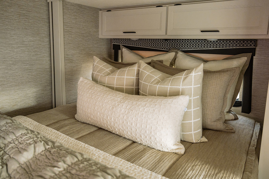 A bed made in an RV with tan woven zipper bedding and an assortment of cream, white and green pillows and blankets.