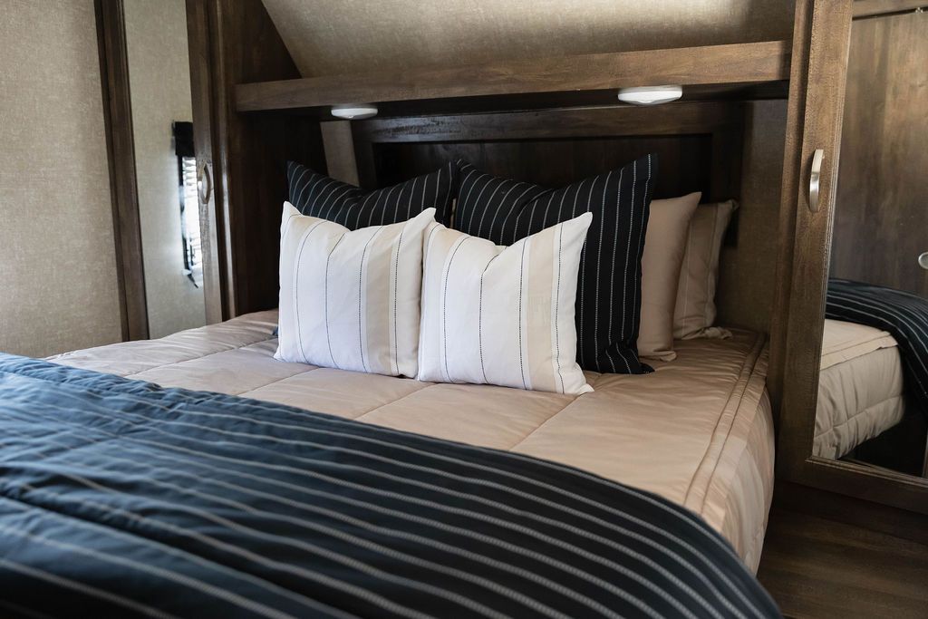 A bed made in an RV with tan zipper bedding and an assortment of neutral and black striped pillows and blanket.