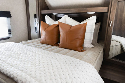A bed made in an RV with khaki green zipper bedding and an assortment of cream and leather pillows and blanket.
