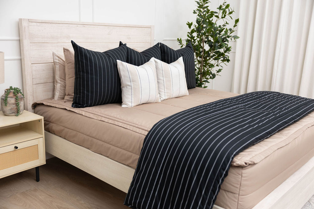 A brightly lit bedroom with a bed made with tan zipper bedding and an assortment of neutral and black striped pillows and blanket.