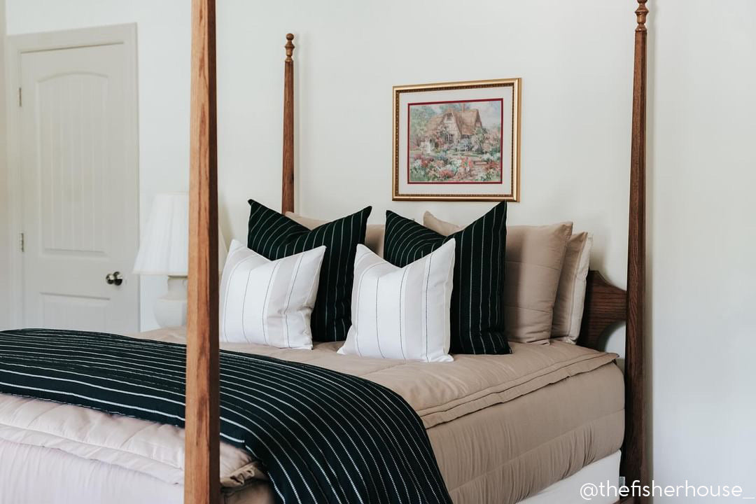 A brightly lit bedroom with a bed made with tan zipper bedding and an assortment of neutral and black striped pillows and blanket with a picture hanging above the bed frame.