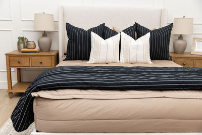 A bed made with tan zipper bedding and an assortment of neutral and black pillows and blanket.