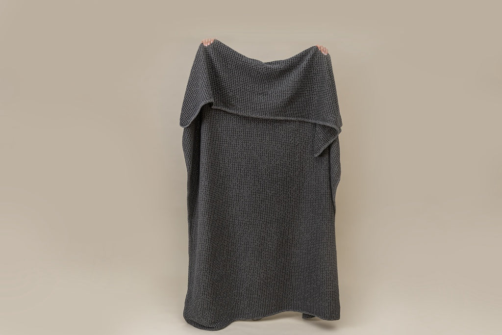 A person holding a  gray textured waffle weave blanket on a tan background.