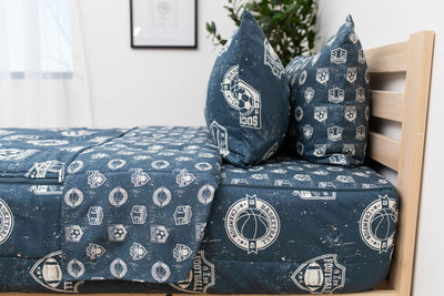A side view of a twin bed made with blue sports themed kids zipper bedding, the top unzipped and pulled back slightly and two pillows at the head of the bed