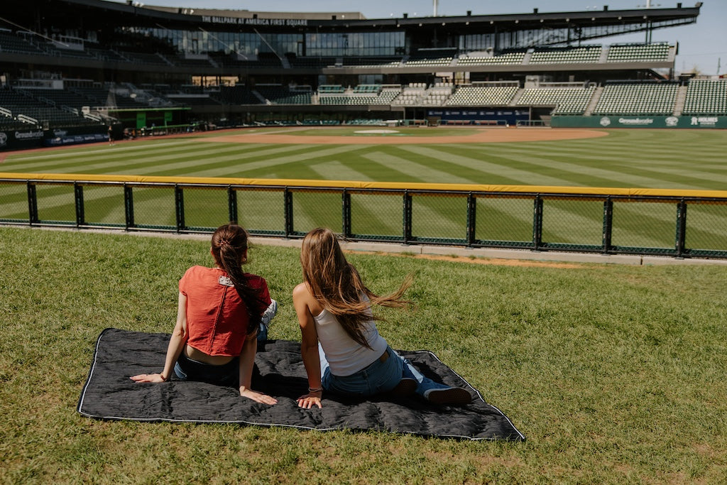 Two women sitting on a grass hill on a water resistant stadium blanket watching a baseball game. 