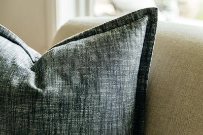 A close up corner view of a large charcoal gray pillow arranged on a gray couch.