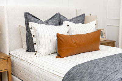 A close up view of a bed made with white and pinstripes zipper bedding and an assortment of neutral and dark charcoal gray pillows and blanket.