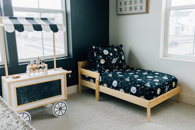 A toddler bed made with black planets and stars kids zipper bedding, a toy serving cart next to the bed and a play tent in the corner.