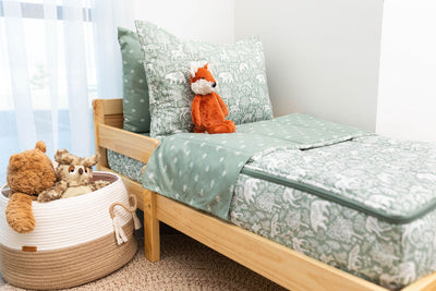 A toddler bed made with sage green forest animal kids zipper bedding and a basket next to it filled with stuffed animals