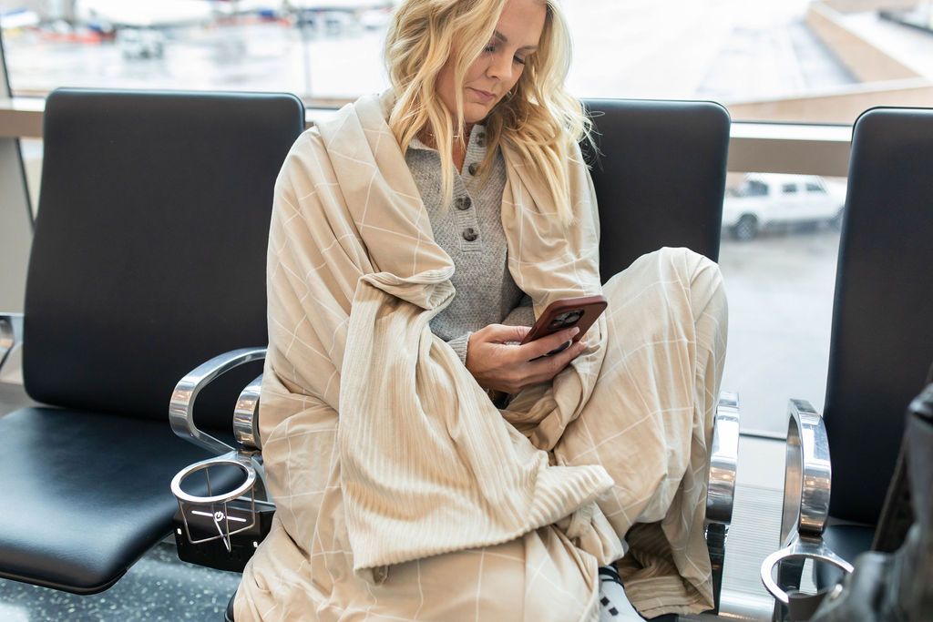 A woman with a light tan grid pattern blanket around her sitting on an airport seat looking at her phone.