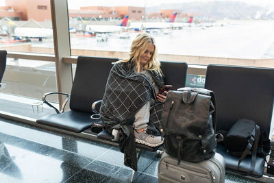 A person with a charcoal gray grid pattern blanket on them sitting on an airport chair with their luggage by them looking at their phone.